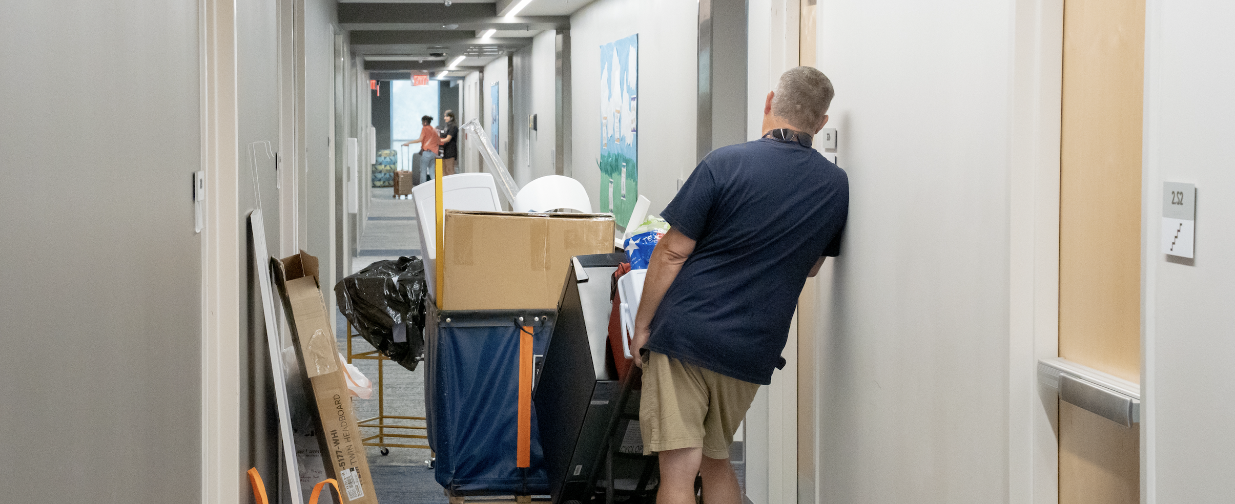Male adult standing with moving dolly loaded with a box with other moving boxes lining a hallway.